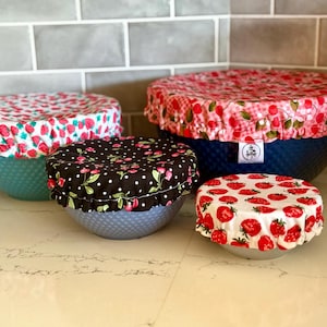 May include: Four round fabric bowl covers with elastic edges, featuring various patterns including red strawberries on white, red cherries on a pink gingham background, and red cherries with green leaves on a black background with white polka dots.