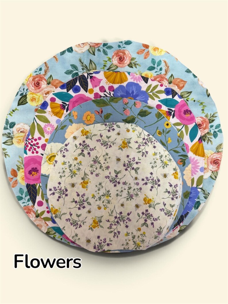 K&ouml;nnte beinhalten: Ein Set aus vier kreisf&ouml;rmigen Sch&uuml;sseldeckeln aus Stoff, die in absteigender Gr&ouml;&szlig;e gestapelt sind. Die Muster zeigen verschiedene florale Designs in Rosa-, Gelb-, Blau- und Gr&uuml;nt&ouml;nen, wobei der kleinste Deckel einen zarten Lavendel- und Bienenaufdruck aufweist. Das Wort Flowers ist unten links in Schwarz gedruckt.