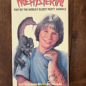 Peut inclure: Une jaquette de cassette VHS pour le film Prehysteria! montrant une personne en chemise bleu clair entourée de trois petits dinosaures. Le texte indique Prehysteria! They're the world's oldest party animals et Plus! Video Bonus-Moonbeam's Videozone.