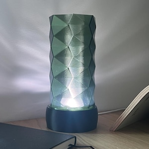 May include: A geometric, green-toned lamp with a cylindrical shape and a diamond pattern. The lamp sits on a dark base and emits a soft, diffused light. A notebook and an open book are visible on the wooden surface.
