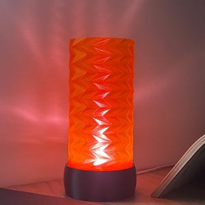 May include: A geometric patterned lamp in shades of orange and yellow, with a dark base. The cylindrical lamp emits a warm, red light. It sits on a wooden surface, next to a closed notebook and an open book.