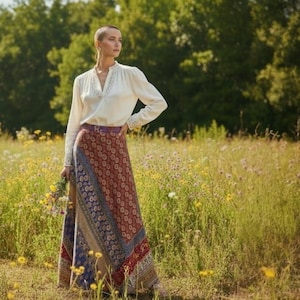Peut inclure: Une longue jupe à motifs dans les tons de rouge, bleu et beige, associée à un chemisier blanc avec des détails en dentelle. La jupe présente un motif patchwork. La scène se déroule dans un champ de fleurs sauvages, créant une esthétique bohème.