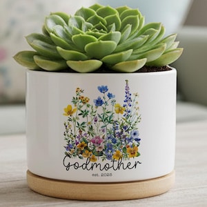 May include: A white ceramic plant pot with a green succulent. The pot has a floral design and the word "Godmother" with "est. 2025". It rests on a round wooden base. The pot is suitable for indoor plants.