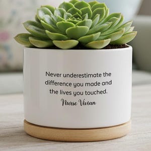 Puede incluir: Una maceta de cerámica blanca con una planta suculenta verde. La maceta tiene el texto "Never underestimate the difference you made and the lives you touched. Nurse Vivian" y se asienta sobre una base de madera.