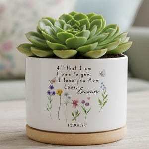 Puede incluir: Una maceta de cerámica blanca con una planta suculenta verde. La maceta tiene una base de madera y presenta un diseño floral con mariposas y el texto "All that I am I owe to you. I love you Mom Love, Emma 11.04.25."