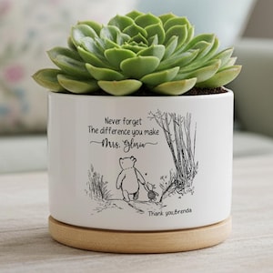 Puede incluir: Maceta de cerámica blanca con una planta suculenta verde. La maceta tiene una base de madera y presenta una ilustración en blanco y negro de un oso y el texto "Never forget the difference you make Mrs. Gloria".