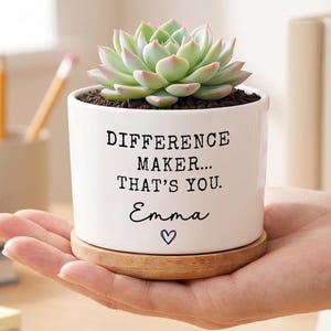Puede incluir: Una maceta de cerámica blanca con una planta suculenta. La maceta tiene el texto "DIFFERENCE MAKER... THAT'S YOU." y el nombre "Emma" con un símbolo de corazón. La maceta está sobre una base de madera.