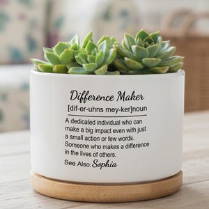 Può includere: Un vaso di ceramica bianca con la scritta "Difference Maker" e una definizione. Il vaso contiene diverse piante grasse verdi e poggia su una base di legno. Il testo include il nome "Sophia".