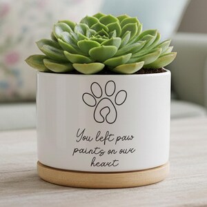 Puede incluir: Una maceta de cerámica blanca con una planta suculenta verde. La maceta tiene un diseño de huella de pata y el texto "You left paw prints on our heart". Se asienta sobre una base redonda de madera.