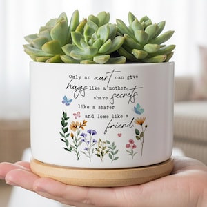 Può includere: Vaso di ceramica bianca con una pianta succulenta, appoggiato su un piattino di legno. Il vaso presenta una citazione con motivi floreali e farfalle. La citazione recita: "Solo una zia può dare abbracci come una madre, condividere segreti come una confidente e amare come un'amica."