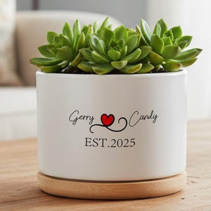 May include: A white ceramic planter with a wooden base, holding green succulents. The planter has the text "Gerry Candy" with a red heart and the year "EST.2025". The planter sits on a wooden surface.