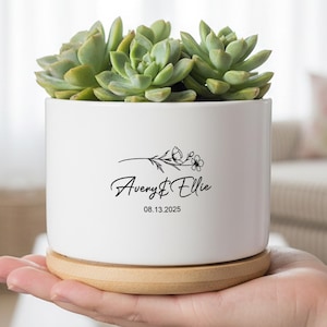 Puede incluir: Una maceta de cerámica blanca con una planta suculenta, sobre un platillo de madera. La maceta tiene los nombres "Avery & Ellie" y la fecha "08.13.2025" en escritura negra. La suculenta tiene hojas verdes.