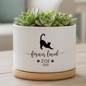 May include: A white ceramic planter with a black cat silhouette, the words "forever loved", a name, and a year. The planter holds several green succulents and sits on a wooden base. A decorative item for the home.