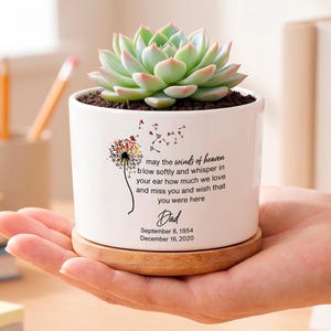 Op de afbeelding: Een witte keramische plantenpot met een bamboe schoteltje en een groene vetplant. De pot is voorzien van een paardenbloemafbeelding en de tekst: may the winds of heaven blow softly and whisper in your ear how much we love and miss you and wish that you were here, Dad, September 8, 1954, December 16, 2020.
