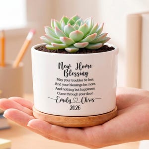May include: A white ceramic planter with a bamboo saucer containing a green succulent. The planter features black text reading New Home Blessing, a poem, the names Emily and Chris with a heart icon, and the year 2026 printed on the front.