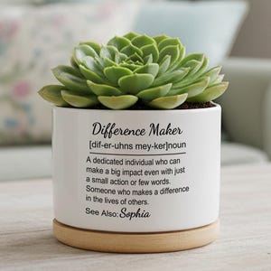 May include: A white ceramic plant pot with a green succulent. The pot features the text "Difference Maker" and a definition in black. It rests on a round, light-coloured wooden base.