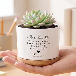 Peut inclure: Un pot en céramique blanc avec une plante succulente, posé sur une soucoupe en bois. Le pot porte l'inscription "Mrs. Smith, THANK YOU FOR BEING A PART OF MY STORY, Emma" en écriture noire. La succulente a des feuilles vertes et roses.