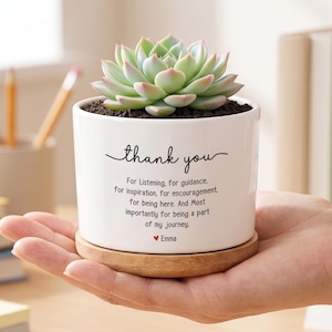 May include: A white ceramic planter with a succulent plant, on a wooden saucer. The planter has the words "thank you" and a message printed in black script. The succulent has green and pink leaves.