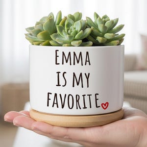 Puede incluir: Una maceta de cerámica blanca con el texto "EMMA IS MY FAVORITE" y un pequeño corazón rojo. La maceta contiene varias suculentas verdes y se asienta sobre un platillo redondo de madera. Una mano sostiene la maceta.