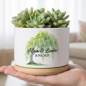 以下が含まれることがあります： 白いセラミック植木鉢、水彩画のしだれ柳のデザイン、緑色の多肉植物入り。 植木鉢には「Klein & Linda」の名前と「JUNE 2025」の日付が記載されています。 植木鉢は丸い木製の台座の上にあります。
