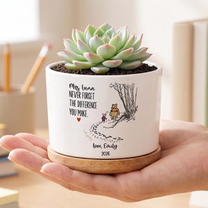 Può includere: Un vaso di ceramica bianco con una pianta succulenta, appoggiato su un piattino di legno. Il vaso presenta un'illustrazione di Winnie the Pooh e il testo "Mrs. Luna, NEVER FORGET THE DIFFERENCE YOU MAKE." Sono inclusi anche un cuore e le parole "love, Emily 2026".