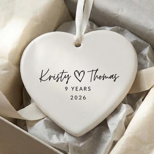 May include: A white heart-shaped ornament with the names "Kristy & Thomas" and the text "9 YEARS 2026" in black script. The ornament has a white ribbon for hanging and is presented in a gift box with tissue paper.