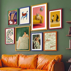 May include: A collection of framed art prints in various sizes and colors, displayed on a green wall above an orange leather sofa. The art features illustrations of people, animals, and abstract designs. The frames are in red, blue, green, and yellow.
