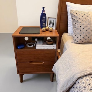 May include: A wooden nightstand with two drawers, an open storage shelf, and a built-in power outlet. The top surface holds a dark tablet, a blue bottle, a small picture frame, and three gold spheres. Two books titled Kinfolk sit on the shelf.
