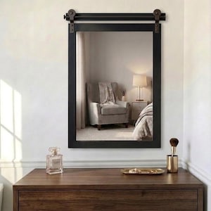 May include: A rectangular wall mirror with a black frame, featuring a decorative metal sliding barn door hardware rail at the top. The mirror is mounted above a dark wood dresser, reflecting a room interior with a chair and a lamp.