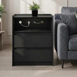 May include: A black nightstand with two drawers and an open top shelf featuring a built-in power outlet with two white cords plugged in. A small green potted plant sits on the top surface of the nightstand, which is positioned next to a gray sofa.