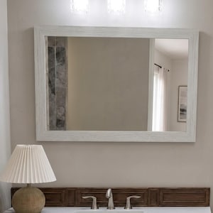 May include: A rectangular wall mirror with a white wood-grain frame mounted above a white bathroom vanity countertop. The vanity features a dark brown wood backsplash, a silver faucet, and a table lamp with a pleated white shade.