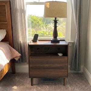 May include: A dark wood nightstand with two drawers, an open storage shelf, and a built-in power outlet. A table lamp with a tan fabric shade sits on the top surface next to a black smartphone charging stand and a small white computer mouse.