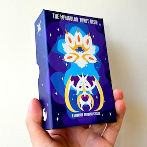 Puede incluir: Una caja de tarot con un diseño celestial. La caja es azul oscuro con una ilustración estilizada de una flor de loto en blanco, naranja y amarillo. El texto "THE LUNISOLAR TAROT DECK" y "A JOURNEY THROUGH CYCLES" están impresos.