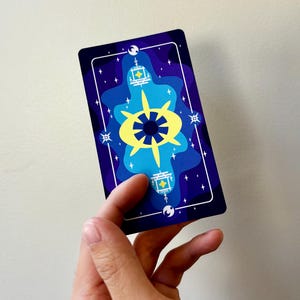 May include: A tarot card with a dark blue background featuring a stylized design in shades of blue and yellow. The design includes a central starburst shape, stars, and geometric elements. The card is held by a hand.