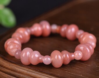 May include: A pink gemstone bracelet featuring a series of gourd-shaped beads and smaller round beads. The bracelet is made of polished, translucent stones, and is displayed on a wooden surface.