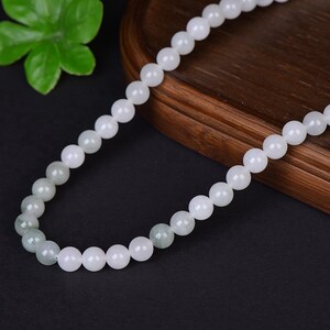 May include: A beaded necklace featuring round, white and pale green beads. The necklace is displayed on a dark wooden surface with a dark background. The beads are strung together, creating a smooth, elegant look. The necklace is a piece of jewelry.