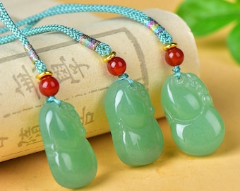May include: Three jade pendants with light green, rounded shapes are displayed. Each pendant is strung on a teal cord with a red bead and gold-colored accents. The pendants are set against a blurred background.