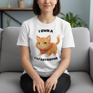 May include: White t-shirt featuring the text "I OWN A catastrophe" and a cartoon orange tabby cat with a red bandana. The cat has green eyes and a smiling expression. The shirt is short-sleeved.