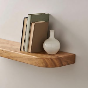 Può includere: Una mensola galleggiante in legno con finitura naturale, che contiene diversi libri e un vaso bianco. La mensola ha un bordo curvo ed è montata su una parete di colore neutro. I libri hanno dorsi di vari colori.