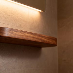 以下が含まれることがあります： 曲がった前面エッジを持つフローティング木製棚。テクスチャ加工された明るい色の壁に取り付けられています。棚は濃い茶色の木材でできており、上部の照明器具で照らされています。 約60cmの長さで、アイテムの展示に最適です。