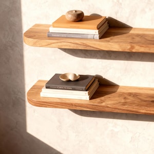 以下が含まれることがあります： 丸みを帯びたエッジの2つの木製フローティングシェルフが、明るい色の壁に取り付けられています。各棚には、本と装飾品が積み重ねられています。棚は明るい茶色の木材でできており、空です。