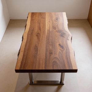 Op de afbeelding: Een rechthoekige houten tafel met een natuurlijke rand en een rijke bruine kleur. De tafel wordt ondersteund door twee gepolijste metalen poten. De afmetingen van de tafel zijn ongeveer 183 cm lang en 91 cm breed.