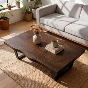 Op de afbeelding: Een donkerbruine houten salontafel met een levendige rand, ondersteund door zwarte metalen poten. Een beige tapijt ligt onder de tafel. Een vaas met gedroogde bloemen, een mok en boeken staan op de tafel. Een grijze bank en planten staan op de achtergrond.