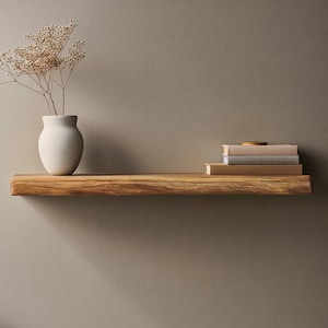 Pode incluir: Uma prateleira flutuante de madeira rústica com borda natural. Um vaso de cerâmica branco com flores secas está à esquerda. Três livros e um pequeno objeto redondo estão empilhados à direita. A prateleira é montada em uma parede de cor neutra.