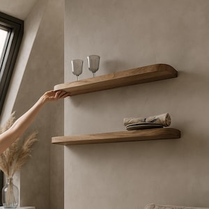 May include: Two wooden floating shelves mounted on a neutral-colored wall. The top shelf holds two gray wine glasses. The bottom shelf displays a rolled-up napkin and a plate. The shelves are made of light brown wood.