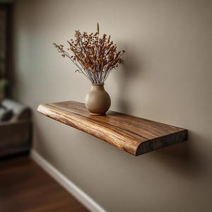 Puede incluir: Estante flotante de madera con borde natural, montado en una pared neutra. Un pequeño jarrón beige contiene flores secas. El estante, de madera oscura, mide unos 75 cm de largo.