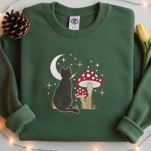 Könnte beinhalten: Dunkelgrünes Sweatshirt mit einem gestickten Design einer schwarzen Katze, einem Halbmond, Pilzen und Sternen. Die Katze sitzt und ist abgewandt. Die Pilze haben rote Kappen mit weißen Punkten. Das Design ist auf der Brust zentriert.