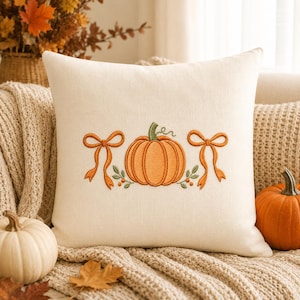 May include: A square off-white throw pillow featuring centered embroidery of an orange pumpkin flanked by two orange bows and small green leafy sprigs. The design is stitched in a simple, clean style on a textured, light-colored fabric cover.