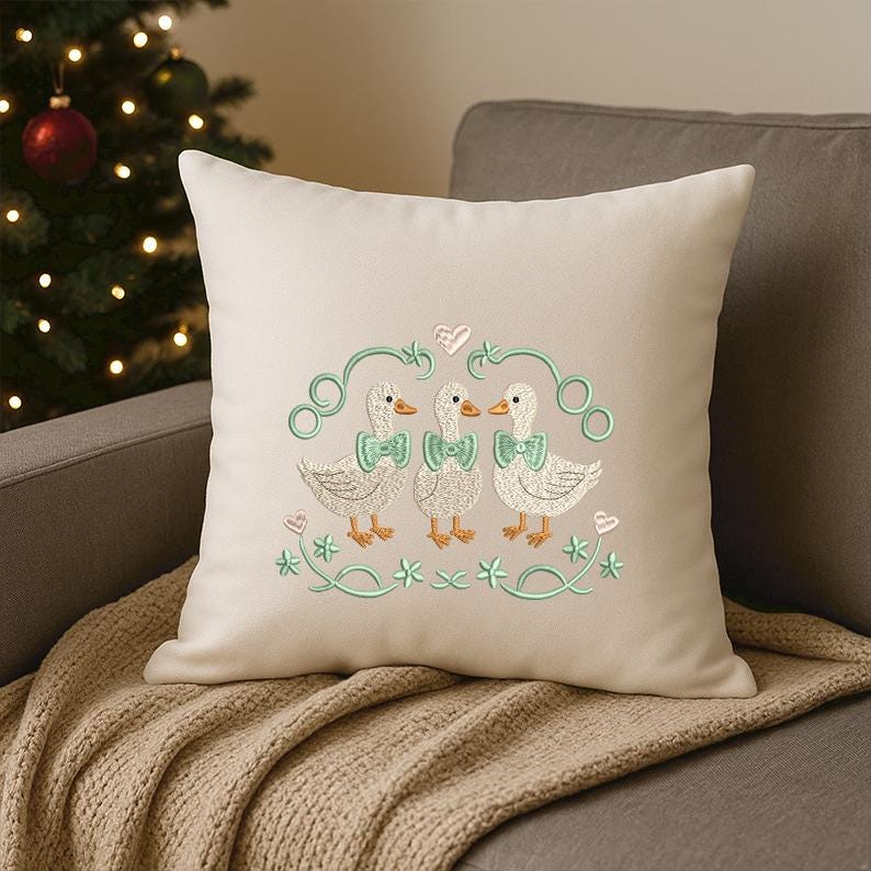 May include: Cream-coloured square cushion with an embroidered design of three geese wearing bow ties. The geese are surrounded by a green floral and heart design. The cushion is on a neutral-toned blanket.