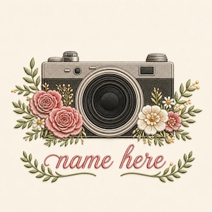 May include: An embroidered design featuring a vintage-style camera in black and gray, surrounded by pink and white floral arrangements with green leaves. Below the camera, the text "name here" is stitched in pink cursive script with leafy accents.